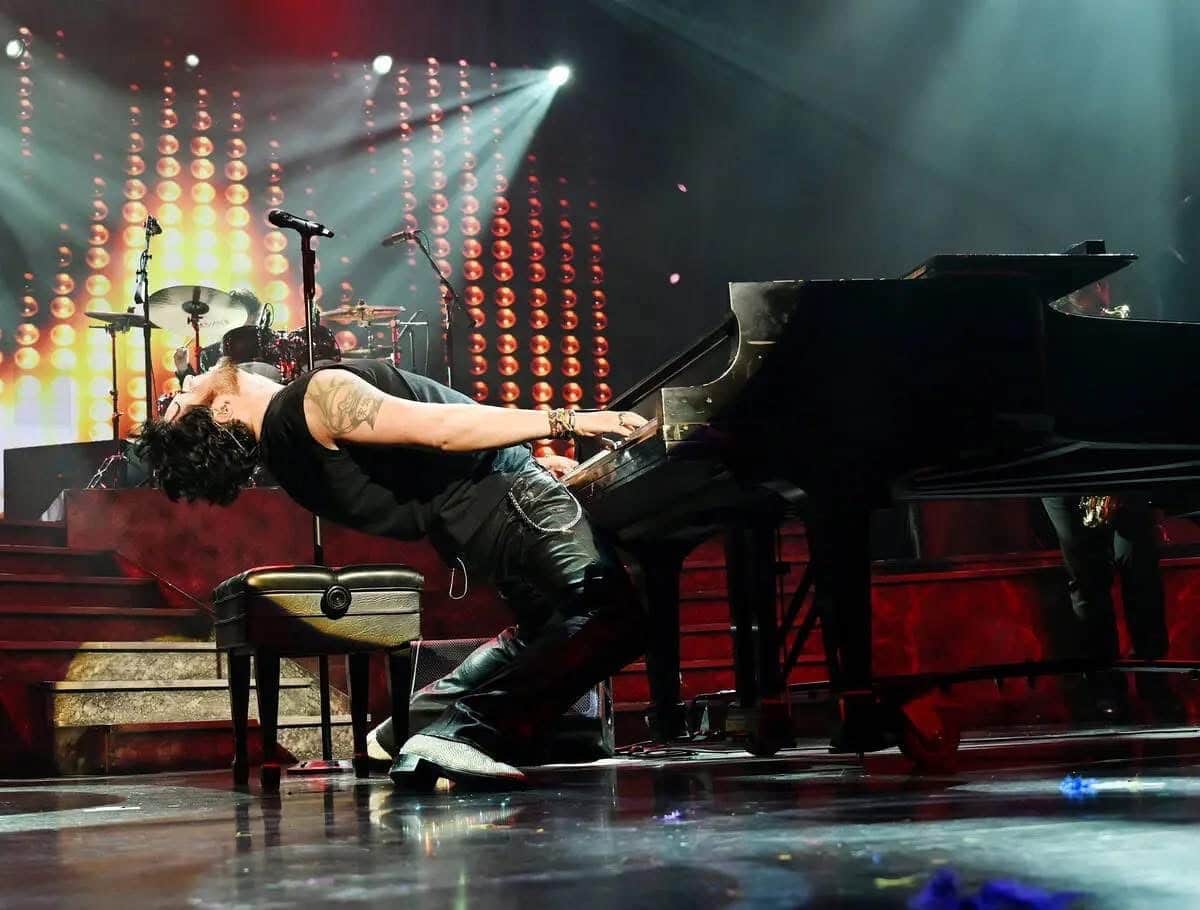 A male musician in a black shirt and leather pants leans back dramatically while playing a grand piano on stage.